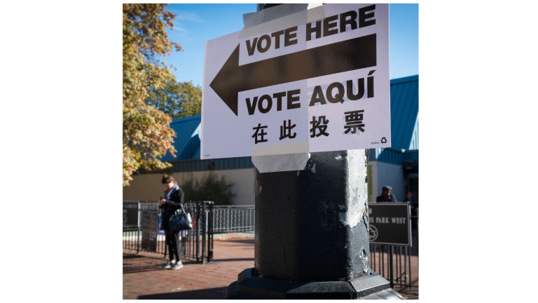 A sign taped to a pole reads Vote Here in English, Spanish, and Chinese, with an arrow pointing left. People stand nearby outside a building, likely a polling station, on a sunny day.