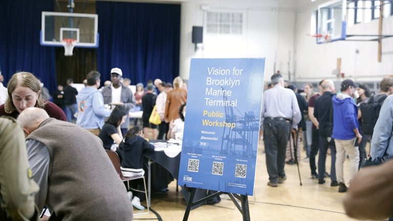 A group of people attend a public workshop in a gymnasium. A blue sign in the foreground reads Vision for Brooklyn Marine Terminal Public Workshop. Tables and informational materials are visible in the background. A group of people attend a public workshop in a gymnasium. A blue sign in the foreground reads Vision for Brooklyn Marine Terminal Public Workshop. Tables and informational materials are visible in the background.