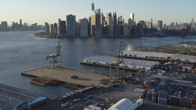 A view of a busy shipping port with cranes and containers in the foreground, and a city skyline with tall buildings across the river in the background under a clear sky. A view of a busy shipping port with cranes and containers in the foreground, and a city skyline with tall buildings across the river in the background under a clear sky.