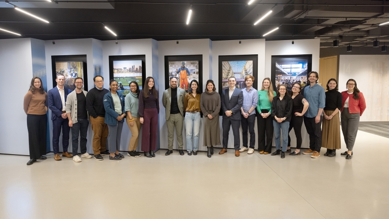 A group of 18 people stand side by side, smiling at the camera in an indoor space, with framed photos of urban and architectural scenes on the wall behind them.