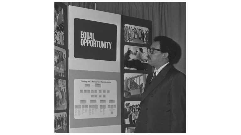 A man in a suit stands in front of a display board with the words “EQUAL OPPORTUNITY,” surrounded by photos of people and an organizational chart labeled “Housing and Development Administration.”.