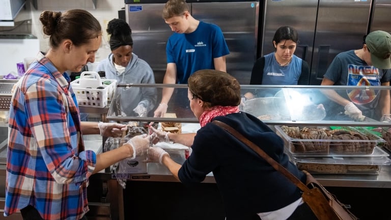 Six people stand behind a counter serving food in a cafeteria or soup kitchen, wearing gloves and focused on preparing and distributing meals. Various trays of food are visible in front of them.
