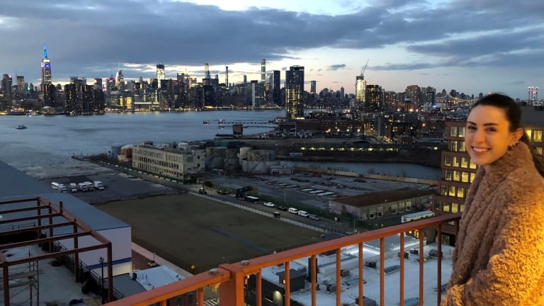 Mary Beth Williams in a brown coat stands on a balcony at sunset, smiling at the camera. Behind her is a New York City skyline with skyscrapers lit up and East River separating the city from the industrial buildings in the foreground. Mary Beth Williams in a brown coat stands on a balcony at sunset, smiling at the camera. Behind her is a New York City skyline with skyscrapers lit up and East River separating the city from the industrial buildings in the foreground.