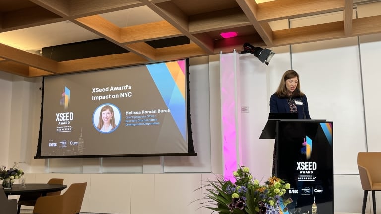 A woman stands at a podium speaking at the XSeed Award event. A large screen behind her displays the title XSeed Award’s Impact on NYC and her name, Melissa Román Burch. Flowers are arranged near the podium. A woman stands at a podium speaking at the XSeed Award event. A large screen behind her displays the title XSeed Award’s Impact on NYC and her name, Melissa Román Burch. Flowers are arranged near the podium.
