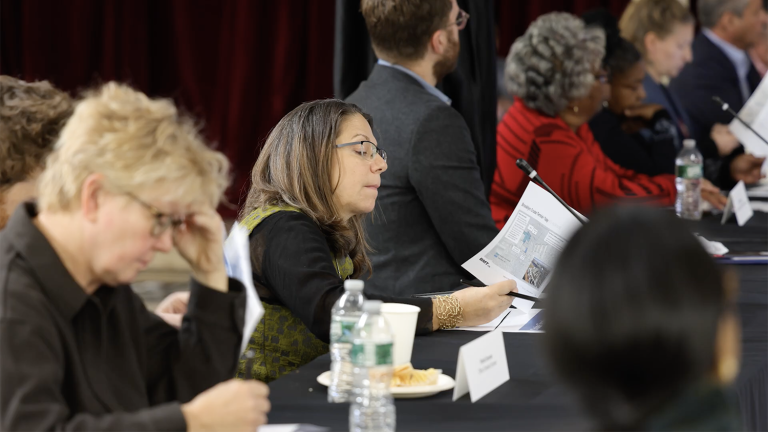 People seated at a long table during a meeting, some reading documents and others listening intently; water bottles, papers, and food are on the table in front of them.
