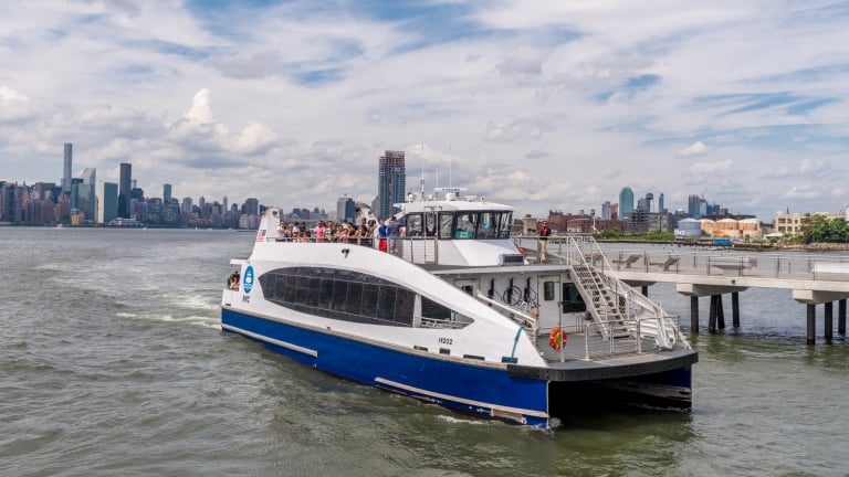 A white and blue NYC ferry boat approaches a dock on the river, with passengers on board and a New York City skyline with tall buildings in the background under a partly cloudy sky.