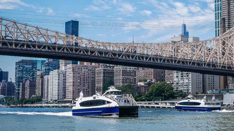 Two blue and white NYC Ferries travel on a river beneath a large steel bridge, with city buildings and skyscrapers in the background under a partly cloudy sky.
