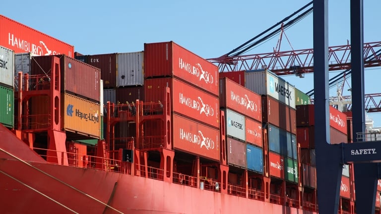 A large red cargo ship loaded with colorful shipping containers is docked at a port, with cranes and blue sky visible in the background. A large red cargo ship loaded with colorful shipping containers is docked at a port, with cranes and blue sky visible in the background.