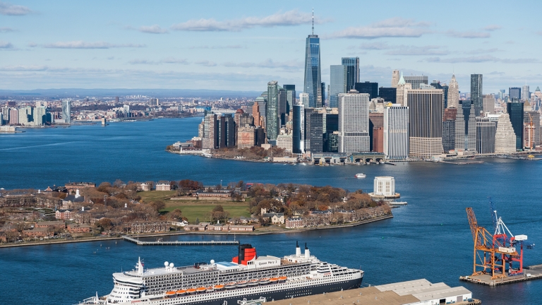 A view of Lower Manhattan’s skyline with tall skyscrapers, the Hudson River, and a large white cruise ship docked at the Brooklyn Cruise Terminal in the foreground near green parks and waterfront piers under a clear sky.