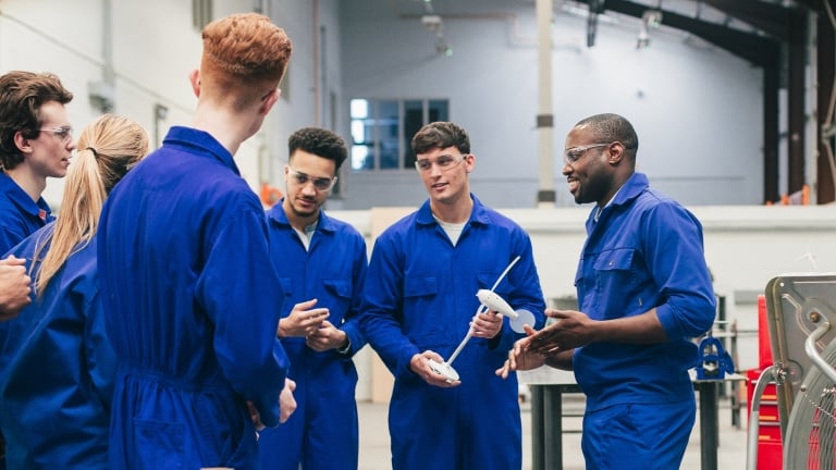 Group of men wearing blue coveralls and safety glasses stand together in a workshop, listening to a man who appears to be explaining something while holding a tool. Industrial equipment is visible in the background. Group of men wearing blue coveralls and safety glasses stand together in a workshop, listening to a man who appears to be explaining something while holding a tool. Industrial equipment is visible in the background.