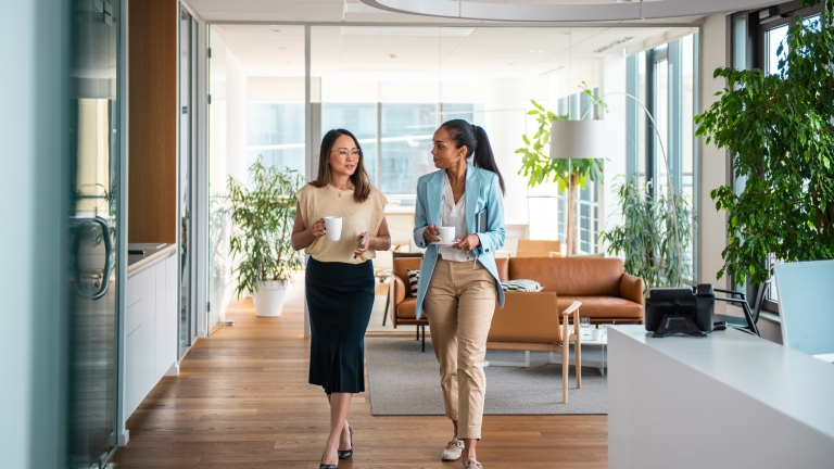 Two women walk and chat in a modern, bright office space with large windows, plants, and a seating area. Both hold coffee mugs and appear to be engaged in a friendly conversation.