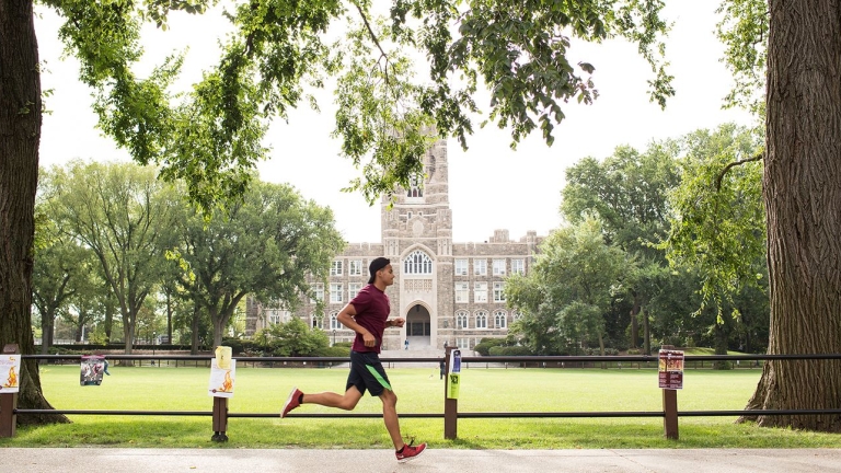 A man in athletic wear jogs on the Fordham University campus along a path lined with trees, with a large, historic stone building and green lawn in the background on a bright, clear day. A man in athletic wear jogs on the Fordham University campus along a path lined with trees, with a large, historic stone building and green lawn in the background on a bright, clear day.