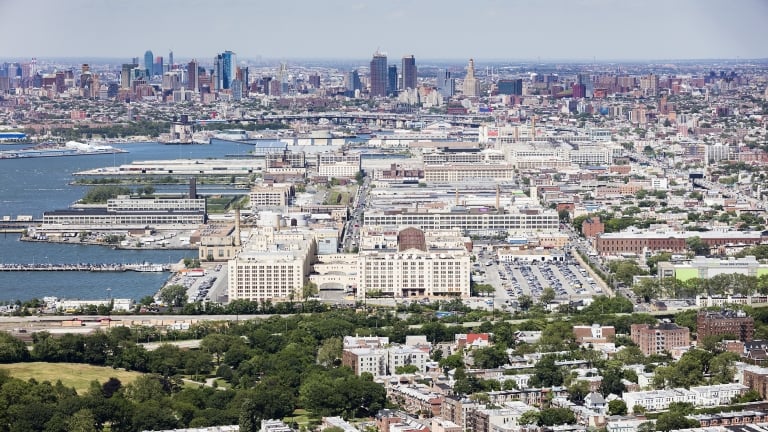 Aerial view of Sunset Park Brooklyn with a mix of residential buildings, green spaces, and waterfront industrial complexes, with a city skyline visible in the distance under a clear sky. Aerial view of Sunset Park Brooklyn with a mix of residential buildings, green spaces, and waterfront industrial complexes, with a city skyline visible in the distance under a clear sky.