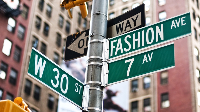 A close-up of street signs at the intersection of Fashion Ave, W 30th St, and 7th Ave in New York City, with tall buildings in the blurred background.