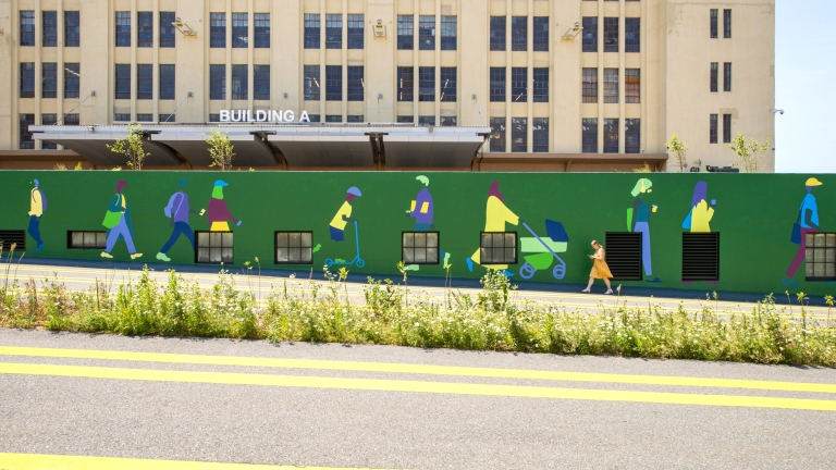 A green mural on a wall depicts colorful silhouettes of people walking and pushing strollers. A person in an orange outfit walks past in front of the mural. Behind the wall is a beige industrial building labeled “Building A.”.