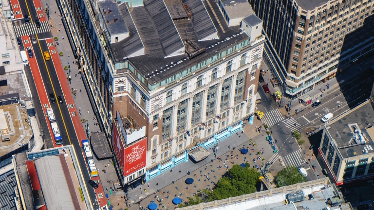 Aerial view of a busy urban intersection with a large building featuring a red sign, pedestrians walking, yellow taxis, and buses on adjacent streets, surrounded by other tall city buildings.