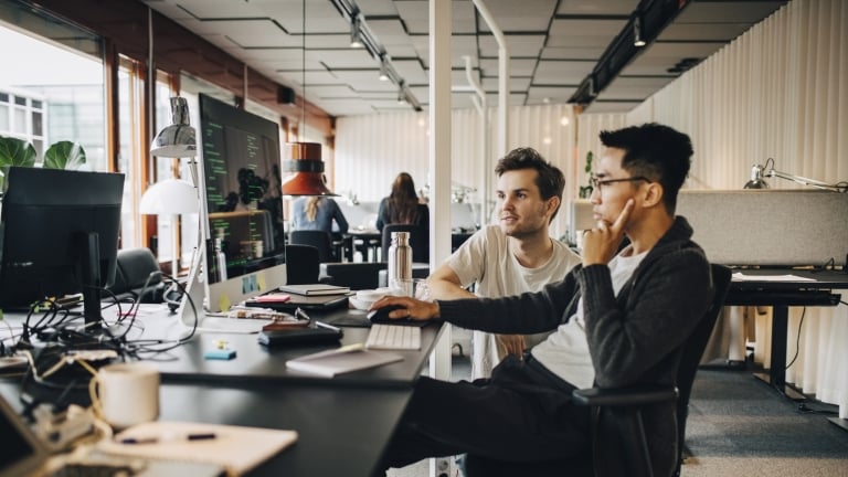 Two men sit at a desk in a modern office, collaborating as they look at a computer screen filled with code. Papers, notebooks, and a water bottle are on the desk, and other people work in the background.