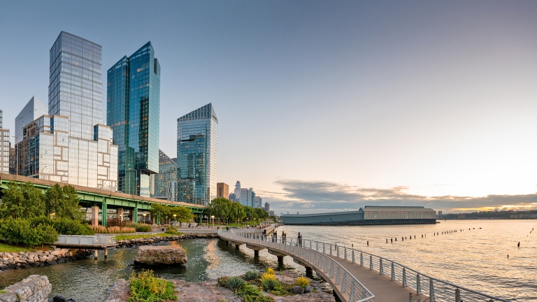 View of the Hudson River Waterfront Greenway, a curving riverside walkway lined with railings and greenery runs alongside modern glass skyscrapers at sunset, with calm water and a distant pier visible on the right.