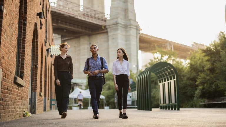 Three people walk and talk together on a city sidewalk near a brick building and a bridge, with trees and a bike rack visible in the background. The scene is bright and appears to be taken during the day.
