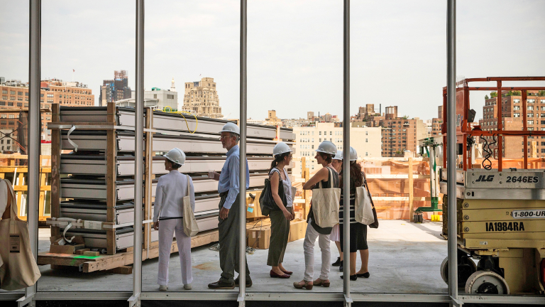 A group of people wearing hard hats stand inside a building under construction, looking out large windows with a city skyline in the background. Construction materials and equipment are visible around them.
