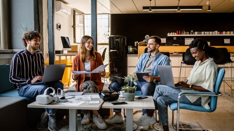 Four people sitting in a modern office, engaged in a casual meeting. They are using laptops and holding papers. The setting includes a coffee table, plants, and a kitchen area in the background, creating a relaxed atmosphere.
