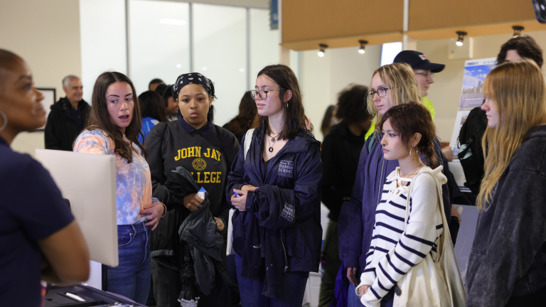A group of young women stand together, listening to a woman at the Maritime Career Fair. Some wear jackets and casual clothes, and one has a “John Jay College” shirt. They appear attentive and engaged with the speaker.