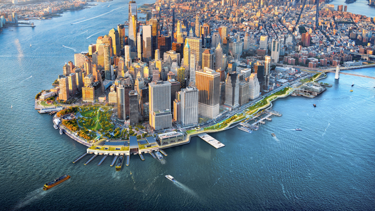 Aerial view of Lower Manhattan, New York City, with tall skyscrapers, surrounding rivers, boats, and the Brooklyn Bridge visible in the background.