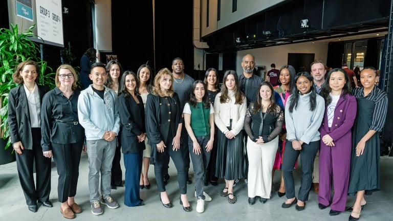 A diverse group of 18 people, both men and women, stand together indoors, posing and smiling for a group photo. They are dressed in business and business-casual attire.