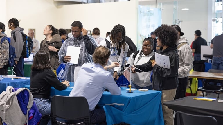 A group of students stands at a table interacting with seated staff at an indoor event. Papers and bags are visible, and other people are seen engaging in the background. The atmosphere appears lively and social.