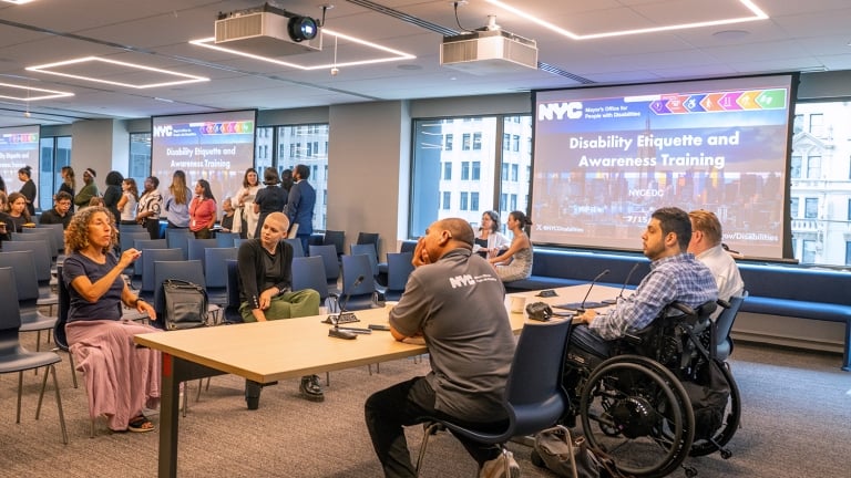 A diverse group of people, including individuals in wheelchairs, converse in a conference room before a presentation titled “Disability Etiquette and Awareness Training” on a projector screen.
