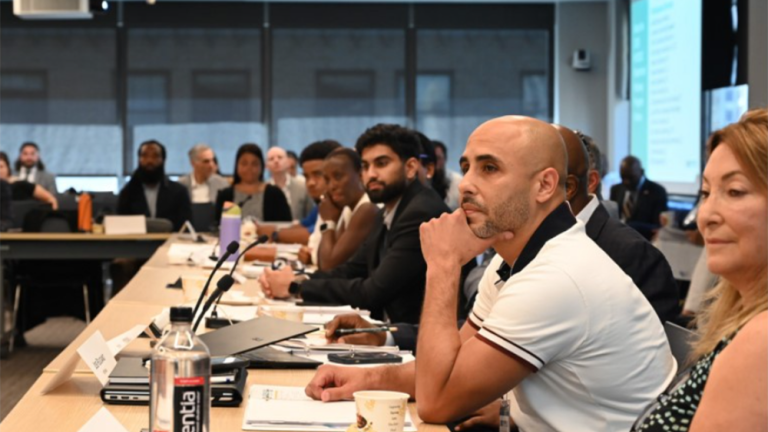 A diverse group of people sit attentively around a large conference table in a modern meeting room, listening to a presentation. Notebooks, cups, and water bottles are visible on the table.
