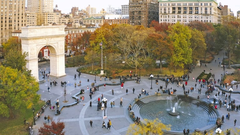 Aerial view of Washington Square Park in New York City with people walking near the arch and around the central fountain, surrounded by autumn trees and city buildings.