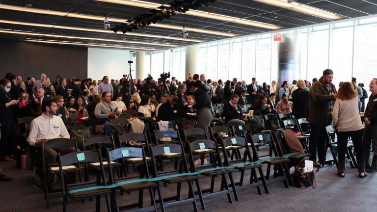 A large group of people gathers in Civic Hall, a modern conference room, with rows of empty chairs in the foreground and bright windows along one wall. Some people are standing, talking, or preparing for the Choose NYC 2025 Summit event to begin.