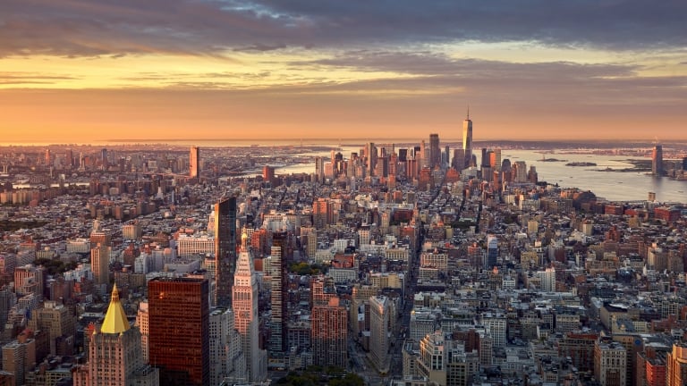 Aerial view of New York City at sunset, with skyscrapers and buildings in the foreground and the sun setting over the Hudson River in the background under a dramatic sky. Photo by Harald Nachtmann.