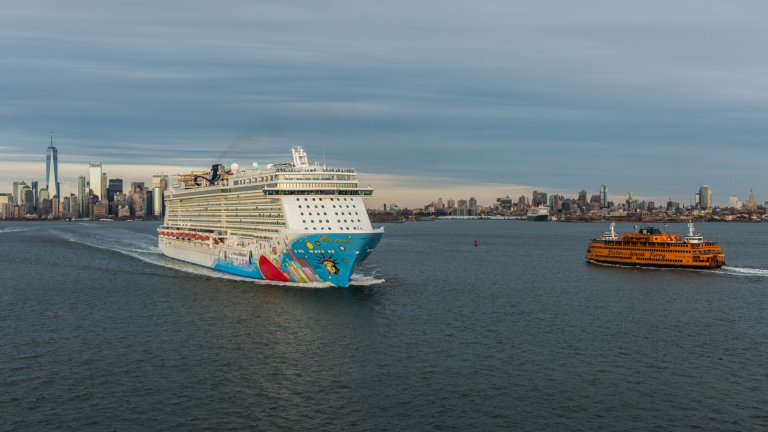 Aerial image of a large, colorful cruise ship and an orange Staten Island Ferry traveling on a river with the New York City skyline, including One World Trade Center, visible under a cloudy sky in the background. Photo courtesy of C. Taylor Crothers.