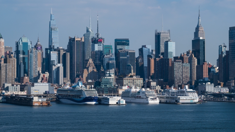 New York City skyline with tall skyscrapers, including the Empire State Building, seen from across the Hudson River with several large cruise ships docked at the Manhattan Cruise Terminal under a blue sky.