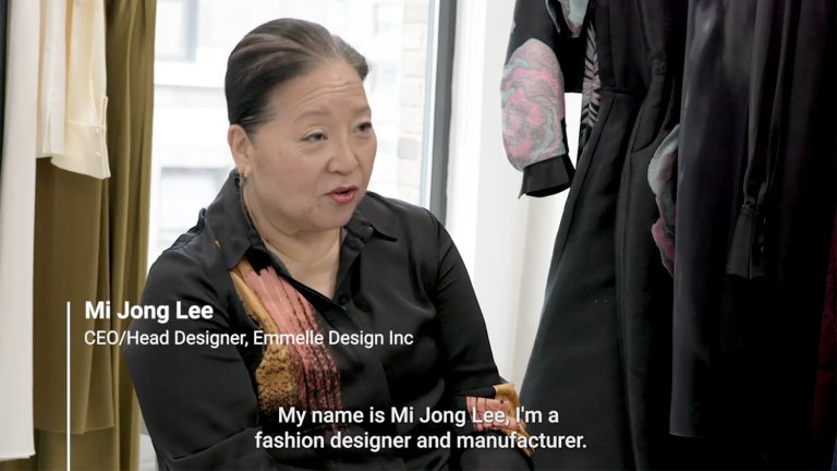 A woman with dark hair tied back, wearing a black shirt with a colorful design, speaks while holding a black garment. On-screen text identifies her as Mi Jong Lee, fashion designer and CEO of Emmelle Design Inc.
