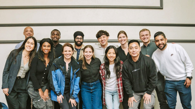 A group of thirteen people stand and kneel together indoors, smiling at the camera. They are dressed casually, and the setting appears to be a social or professional gathering.