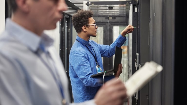 Two IT professionals work in a server room; one in focus adjusts hardware on a server rack while holding a laptop, while the other, blurred in the foreground, inspects a device or document.