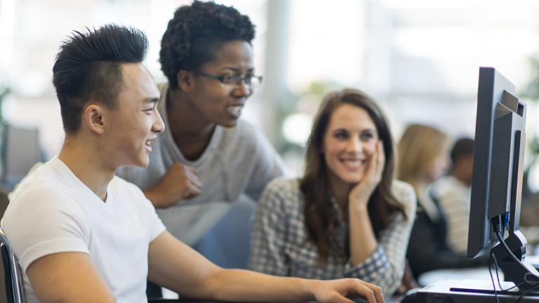 Three young adults work together at a computer. One person types while the others watch and smile, appearing engaged and collaborative in a bright, modern setting.