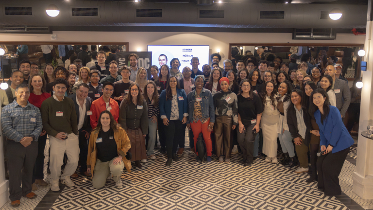 2026 Founder Fellowship cohort pose and smile for a group photo in a well-lit indoor space with patterned floors, mirrors, and a screen in the background. Most of them are wearing name tags.