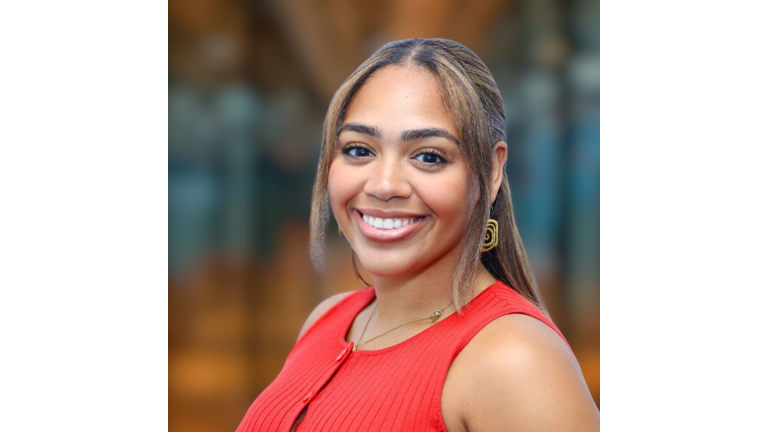 Alexandra Martinez with long, straight hair and a bright smile is wearing a sleeveless red top and gold earrings. Background is softly blurred with warm tones.