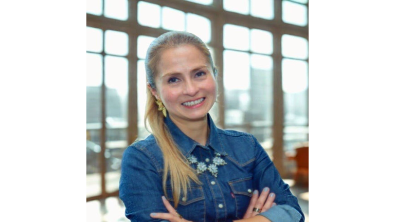 Diana Franco with her long blonde hair in a ponytail, wearing a denim shirt and a statement necklace. She is smiling with her arms crossed in a bright indoor space, with large windows in the background.