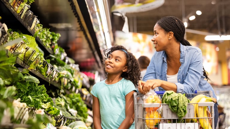 A woman and a young girl smile while shopping for fresh produce in a grocery store. The shopping cart is filled with fruits, vegetables, and groceries, and they are standing near the leafy greens section.