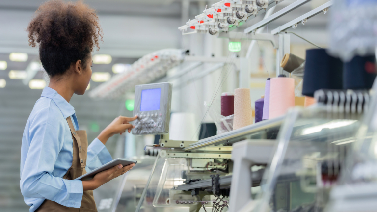 A woman wearing a blue shirt and apron operates a control panel in a modern textile factory, holding a digital tablet beside spools of colorful thread and automated machinery.