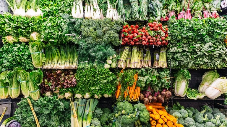 A colorful display of fresh vegetables stacked on shelves, including leafy greens, bok choy, leeks, broccoli, carrots, beets, and lettuce, in a grocery store or market produce section.