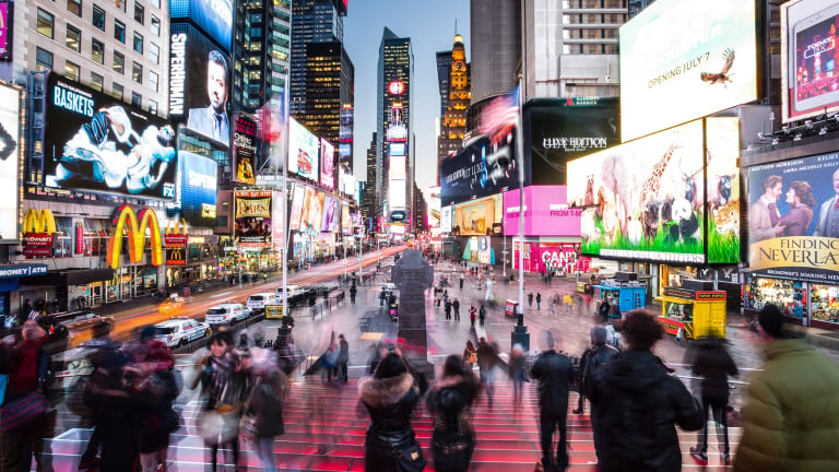 A bustling scene in Times Square, New York City, with blurred crowds, bright lights, colorful billboards, and advertisements illuminating the area at dusk.