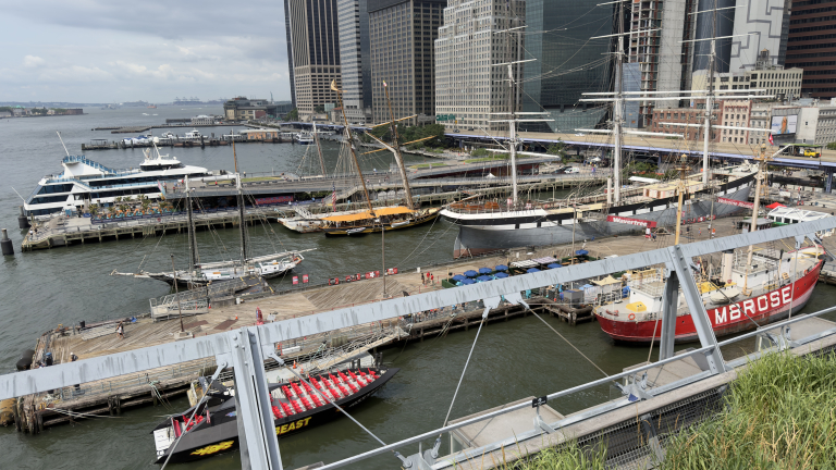 Several historic and modern boats docked at a New York City waterfront, with tall buildings in the background and people walking along the piers.
