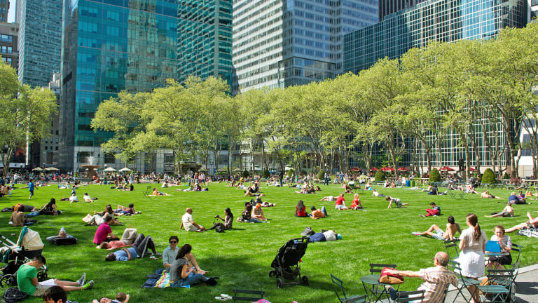 People relax and sit on the grass and in chairs in Byant Park surrounded by tall glass buildings and trees on a sunny day.