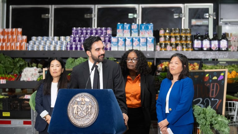 Mayor Mamdani speaks at a podium with the New York City seal in a grocery store with produce and products on shelves behind them.